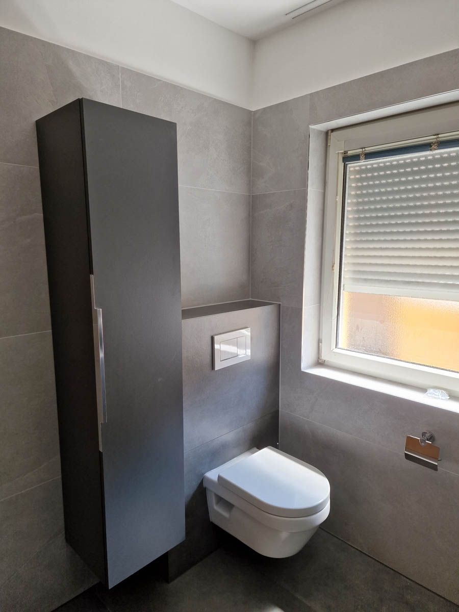 Graues Badezimmer mit wandhängender Toilette, Abstellschrank und Fenster mit Jalousien.