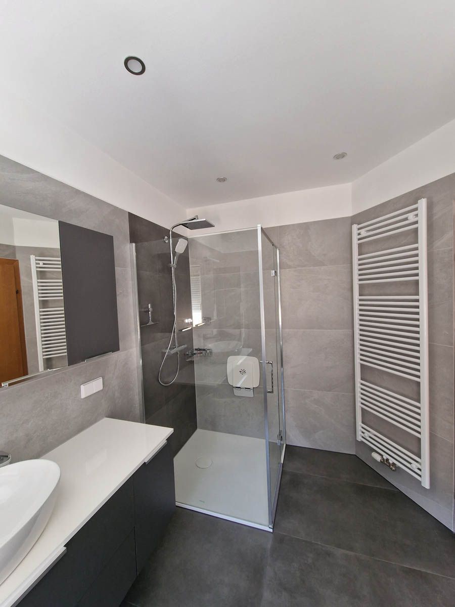 Modernes Badezimmer mit grau gefliesten Wänden, Glasdusche, weißem Waschbecken und Heizkörper.