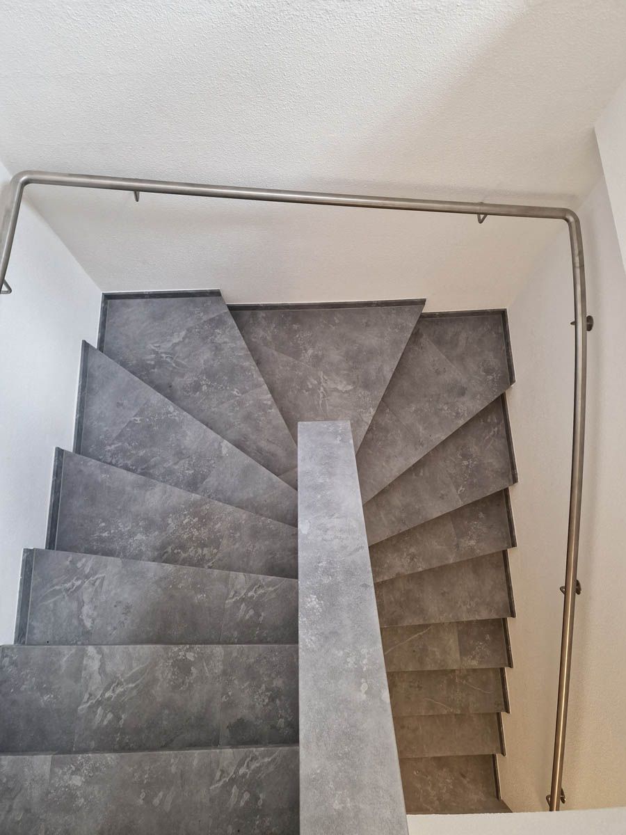 Treppe mit grauen Stufen und einem Metallhandlauf, der von einem weißen Treppenabsatz nach unten führt.