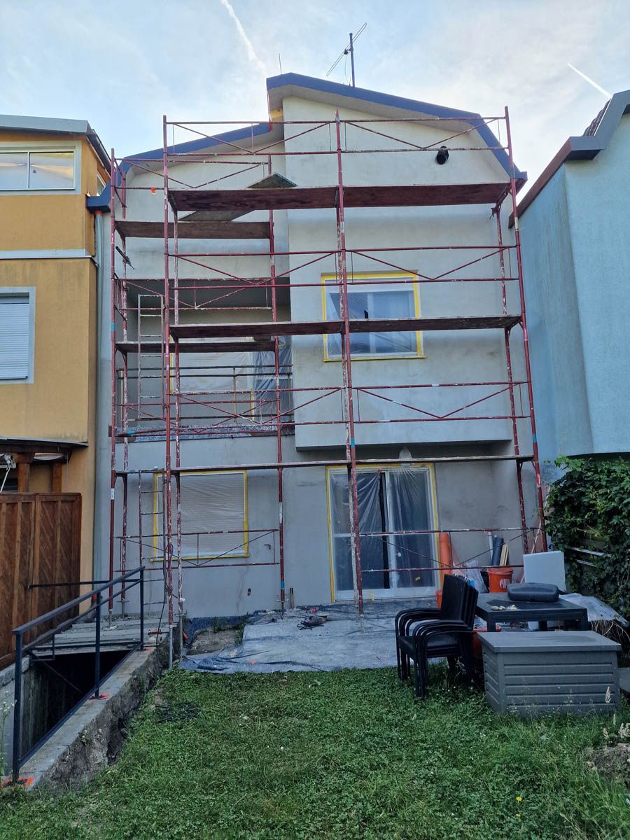Haus im Bau mit Gerüst; hellgrauer Stuck, gelb abgeklebte Fenster und grünes Gras davor.