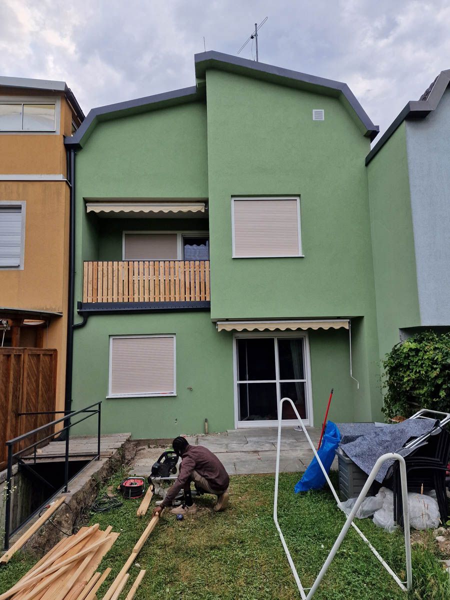 Person sägt Holz in einem Hinterhof. Gewächshaus mit hellbraunen Markisen und Balkon, angrenzend an andere Gebäude.