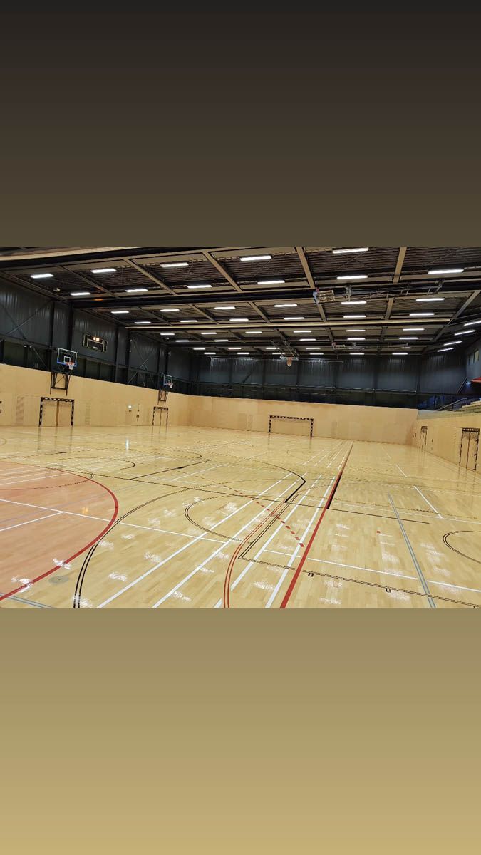 Indoor-Sporthalle mit Holzboden, der für verschiedene Spiele markiert ist. Schwarzes Netz umgibt den Raum.
