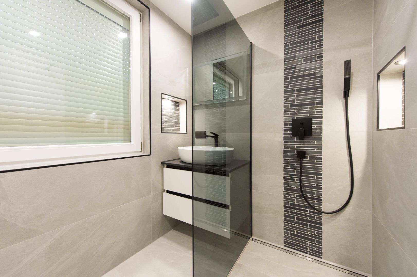 Modernes Badezimmer mit grauen Fliesen, Glasdusche, weißem Waschbecken und dunklen Armaturen.