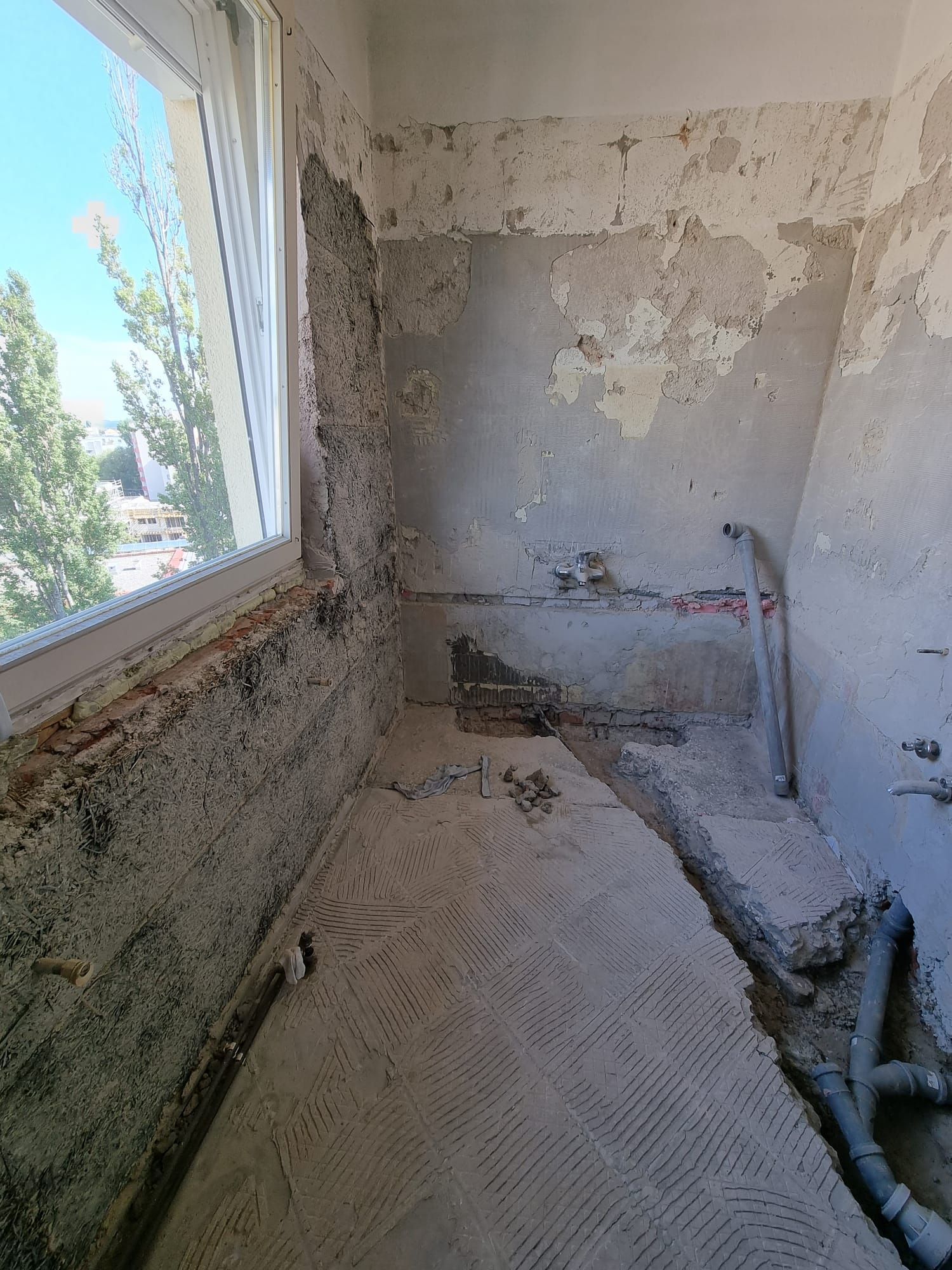 Badezimmerrenovierung im Gange; Wände bis auf die Ziegel freigelegt, freiliegende Rohrleitungen, strukturierter Boden, Fenster mit Aussicht.