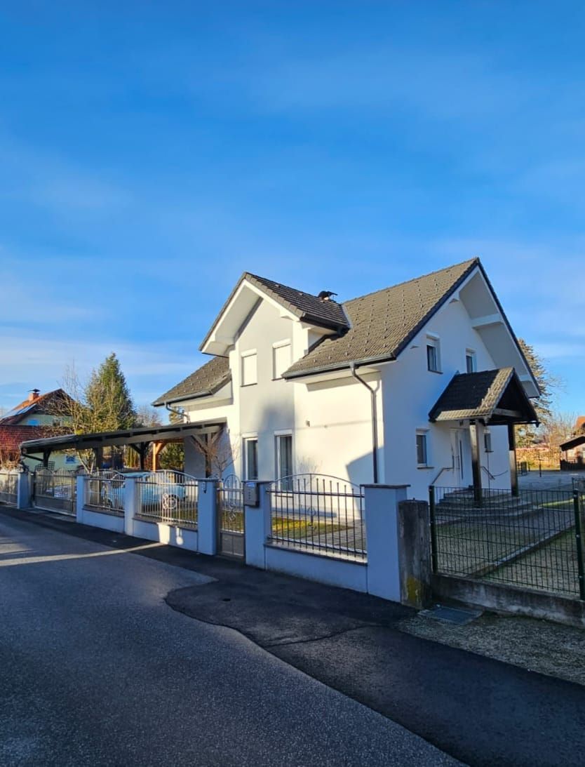 Zweistöckiges hellgraues Haus mit dunklem Dach, hinter einem grauen Zaun, unter einem blauen Himmel.