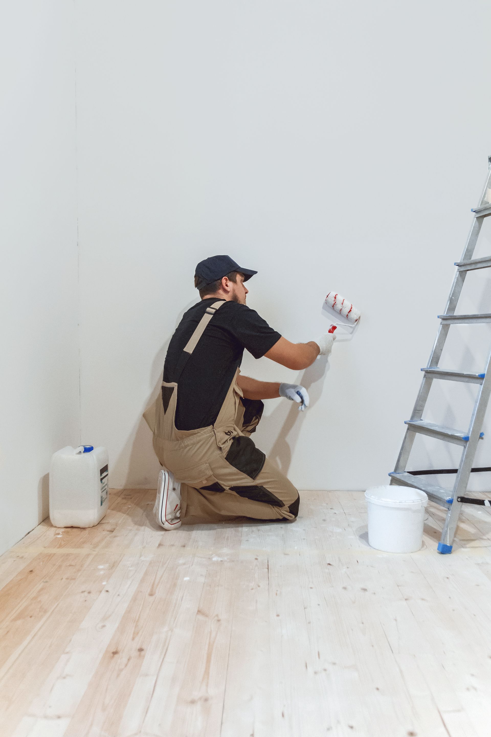Ein Mann in Arbeitskleidung streicht mit einer Rolle eine weiße Wand und steht auf einem Holzboden in einem Raum.