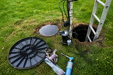 Open utility hole with ladder, cables, and removed round cover on a grassy lawn