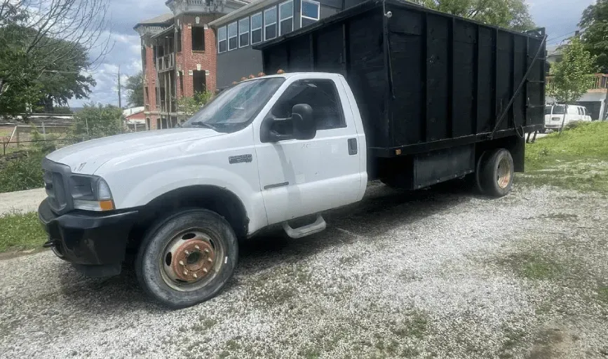 White Ford truck with a black dumpster on a gravel lot.