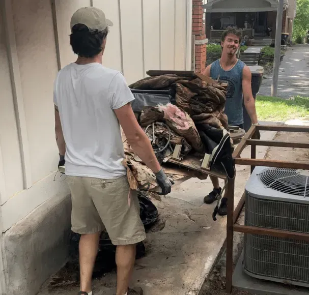 Two men carry debris on a hand truck next to a building and air conditioner.