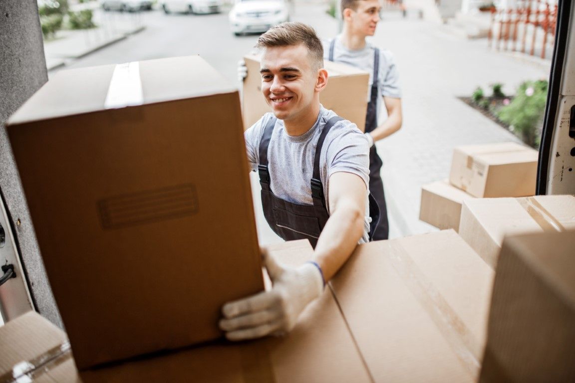 Two men in work clothes loading cardboard boxes into a van, smiling.