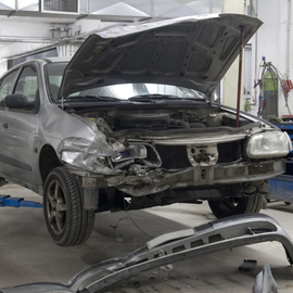 Damaged silver car in auto body shop with hood open and detached front bumper on floor