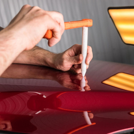 Hands using a white pen-like tool to inspect a glossy red car surface with reflected lights