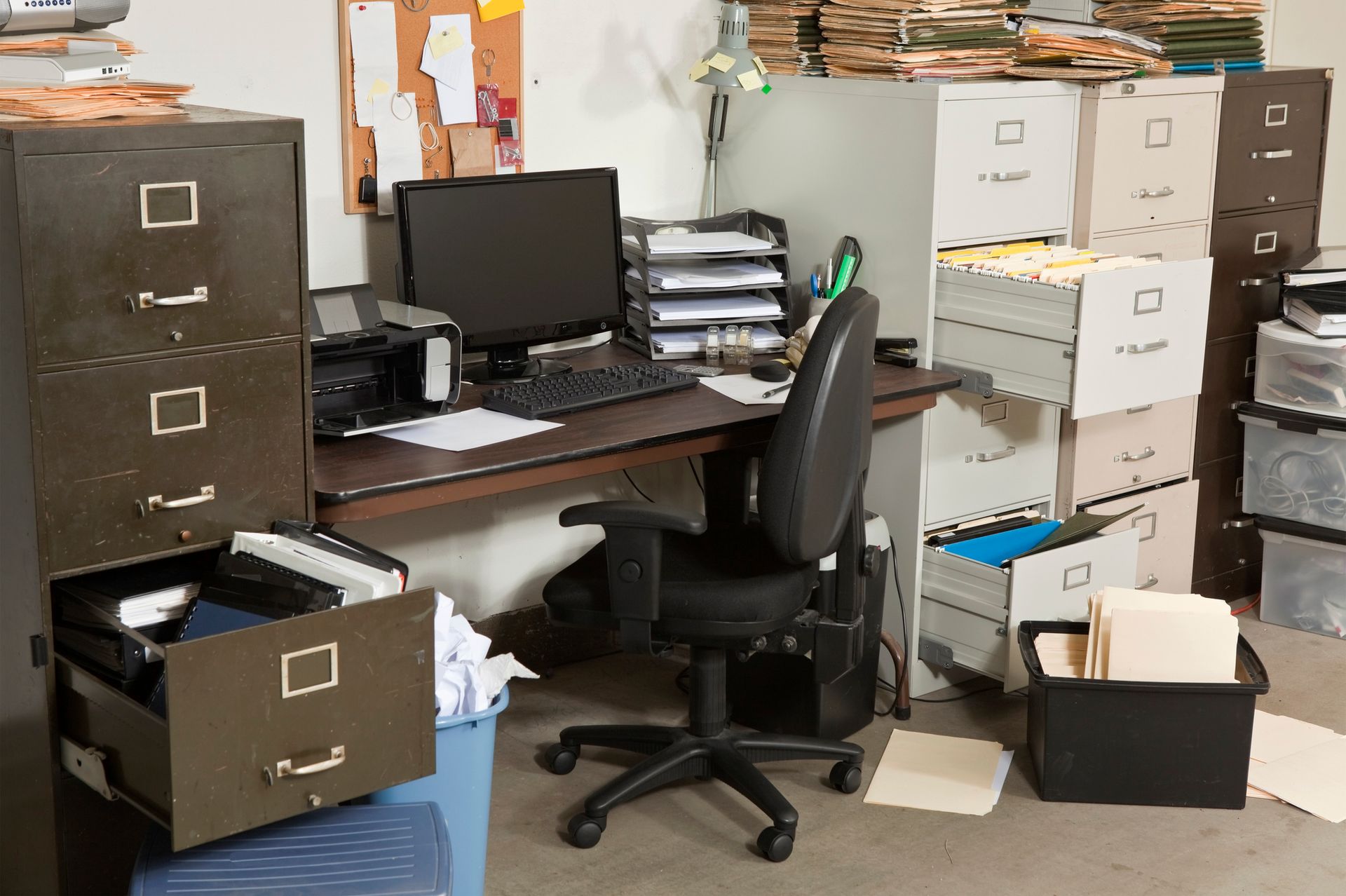 Cluttered home office: metal filing cabinets, computer, stacks of files, and overflowing boxes create a disorganized workspace.