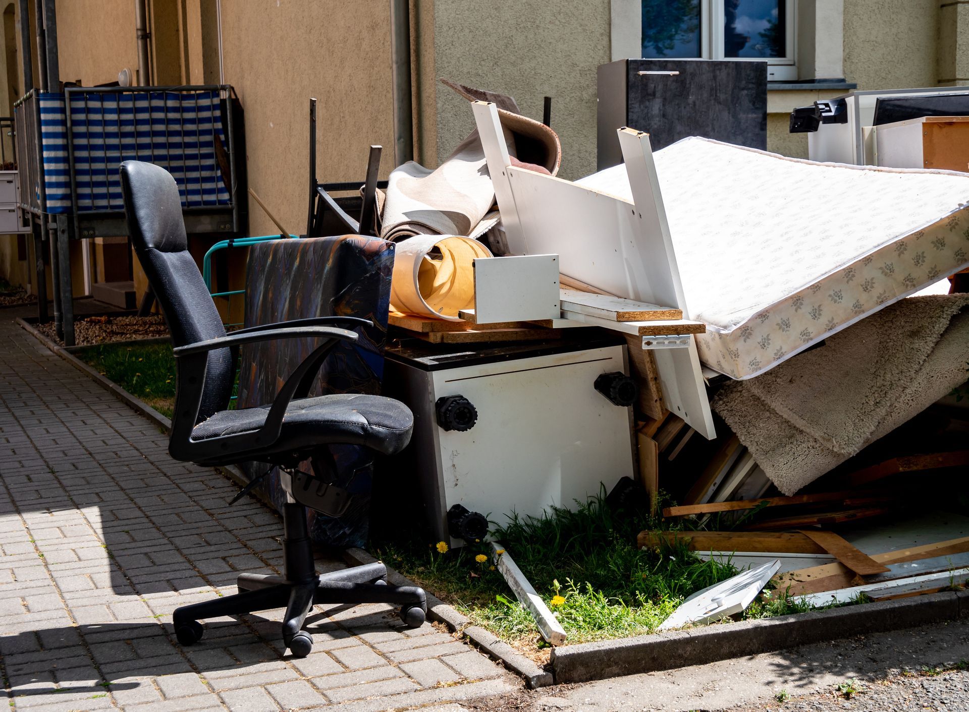 An office chair sits in front of a pile of discarded furniture, including a mattress and appliances, outdoors.