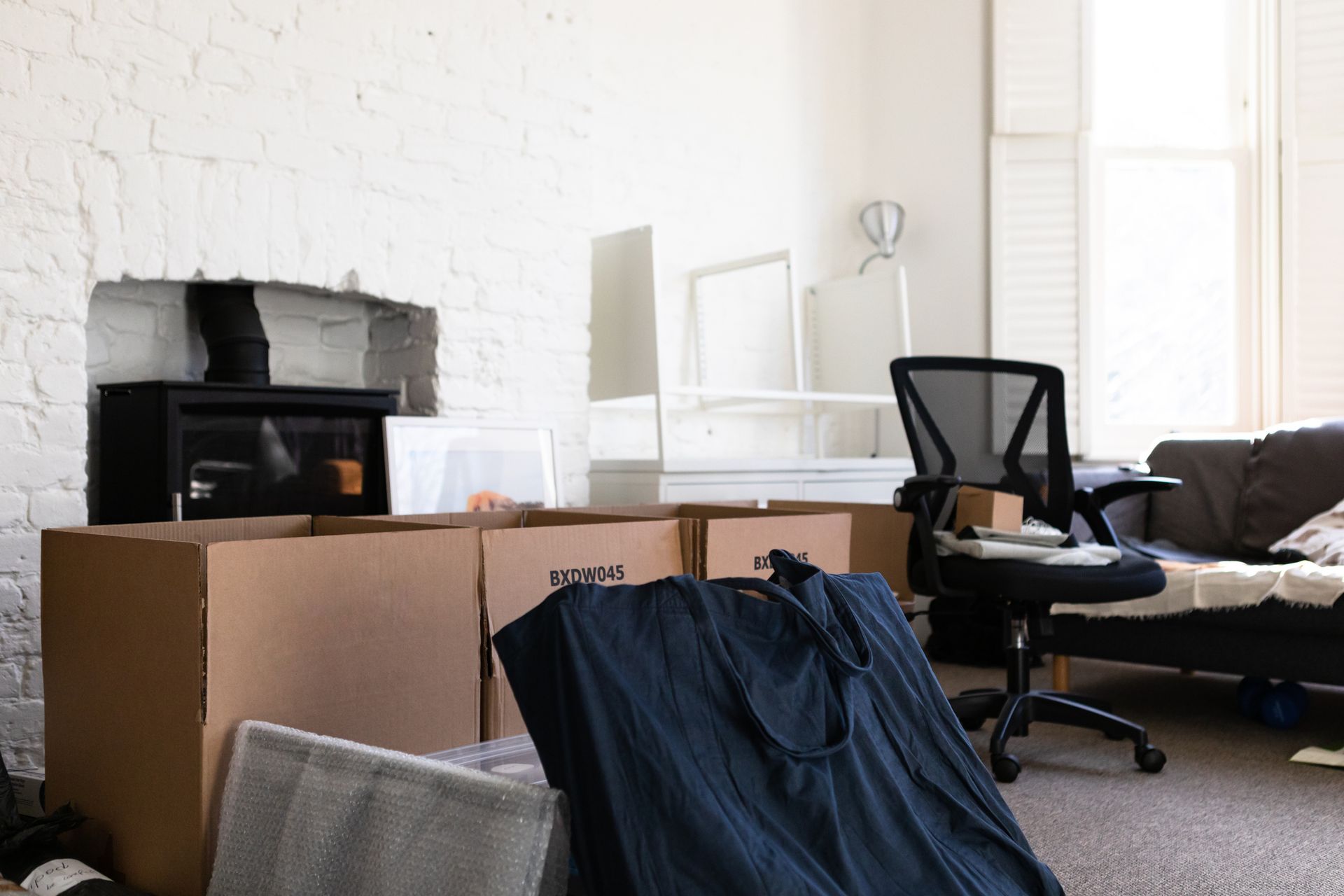 An interior shot showing a room with packed moving boxes, furniture, and a fireplace. A dark chair is in the room.