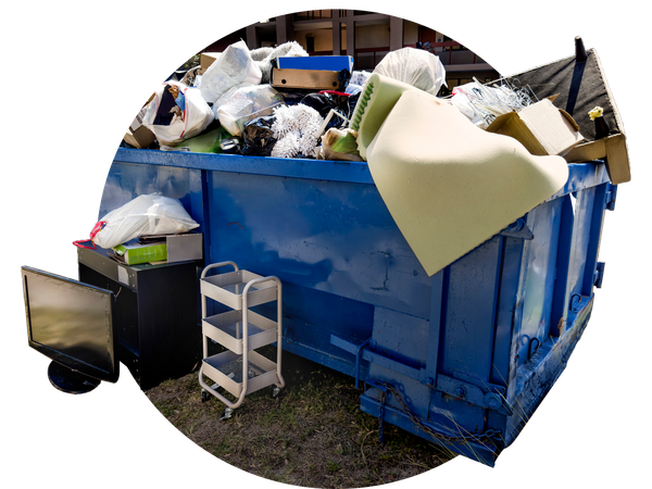 Blue dumpster overflowing with trash, including furniture, cardboard boxes, and a computer monitor, outdoors.