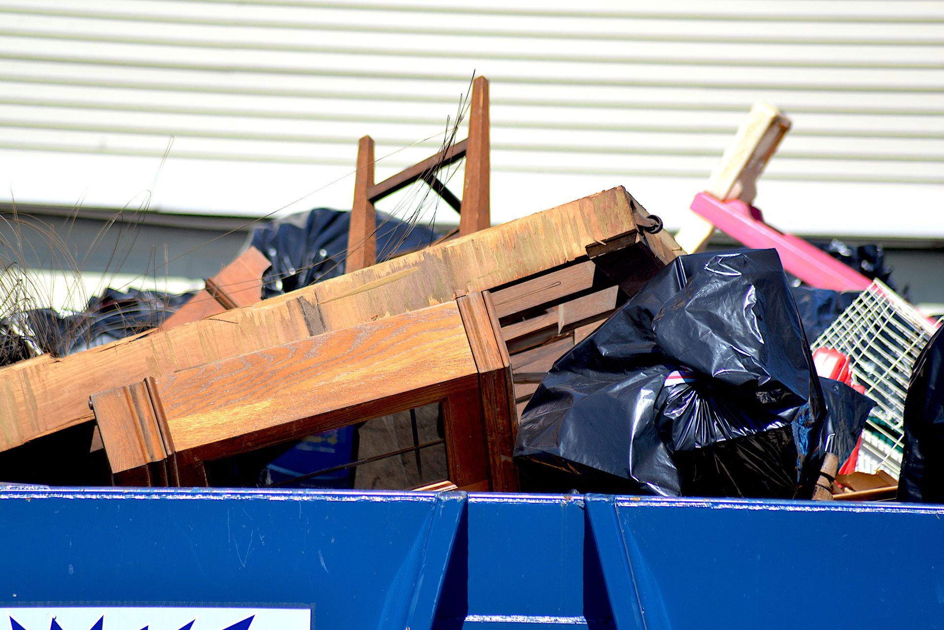 A full blue dumpster overflowing with garbage bags, wooden furniture, and other trash, with a building in the background.
