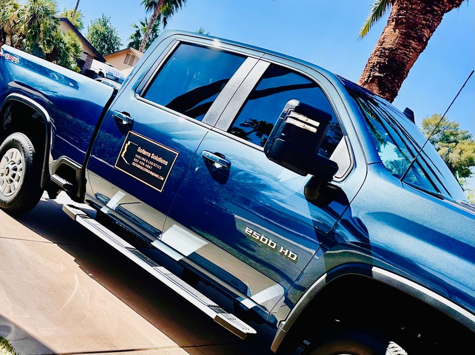 Dark blue Chevrolet Silverado pickup truck parked in a driveway, with the company logo on the door. Palm trees and houses in the background.