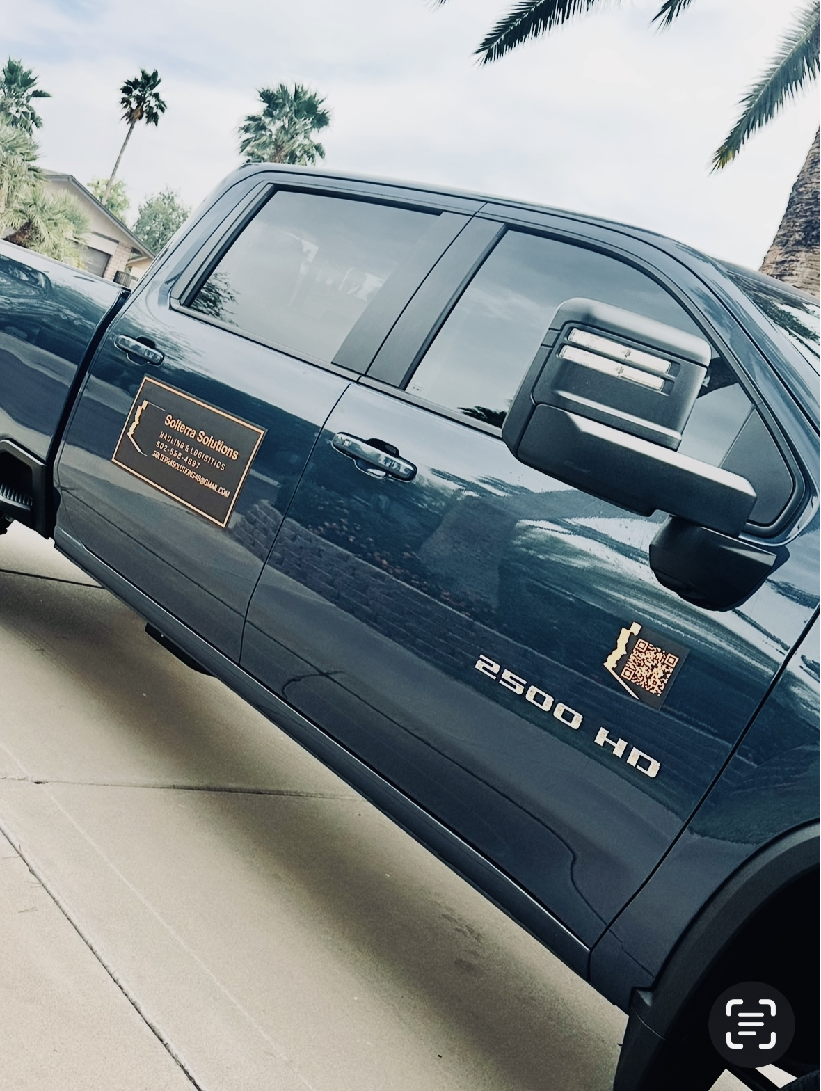 Dark blue Chevy Silverado 2500 HD truck parked on a driveway. Palm trees are visible in the background, and the truck has a company logo on the side.