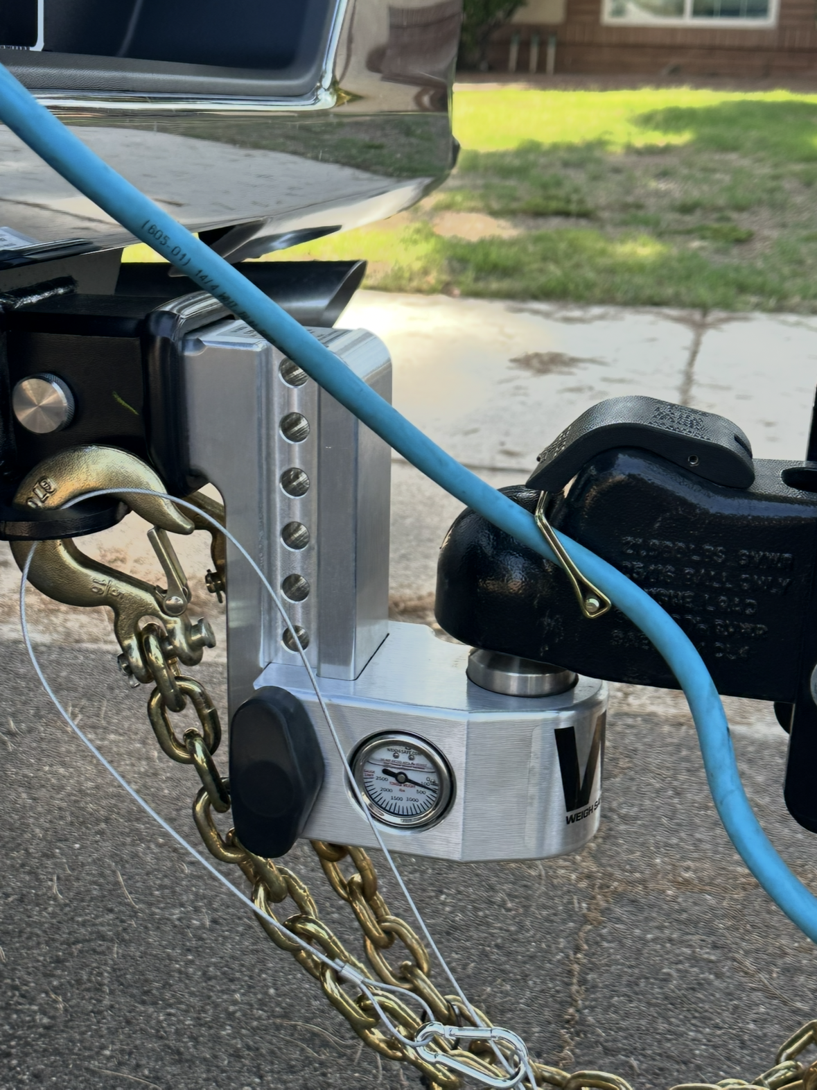 A close-up of a weight distribution hitch attached to a truck. The hitch is silver with a pressure gauge, safety chains, and a blue cable.