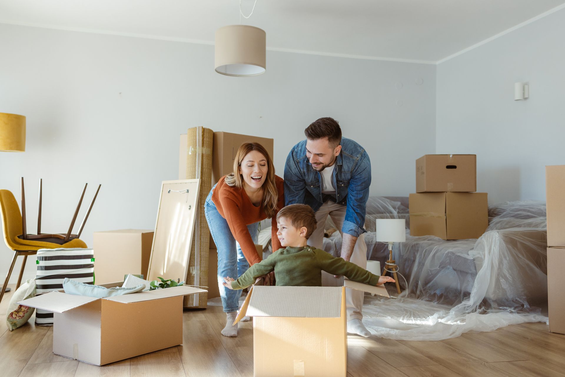 Family unpacking in a new home; son in box pretending to fly, parents smile. Boxes and furniture.