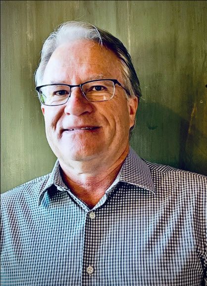 Man with glasses, wearing a button-down shirt, smiles at the camera. Green background.