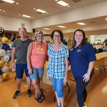 Four people smiling in a brightly lit room with balloons and tables, possibly at an event.