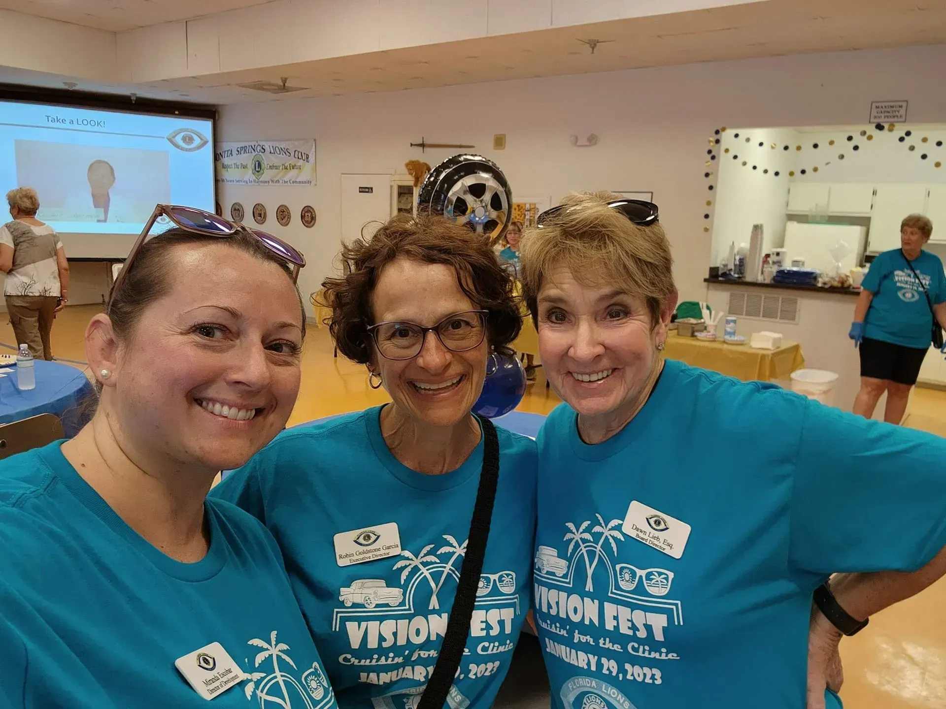 Three women in matching teal shirts smile, posing for a photo at a Vision Fest event.
