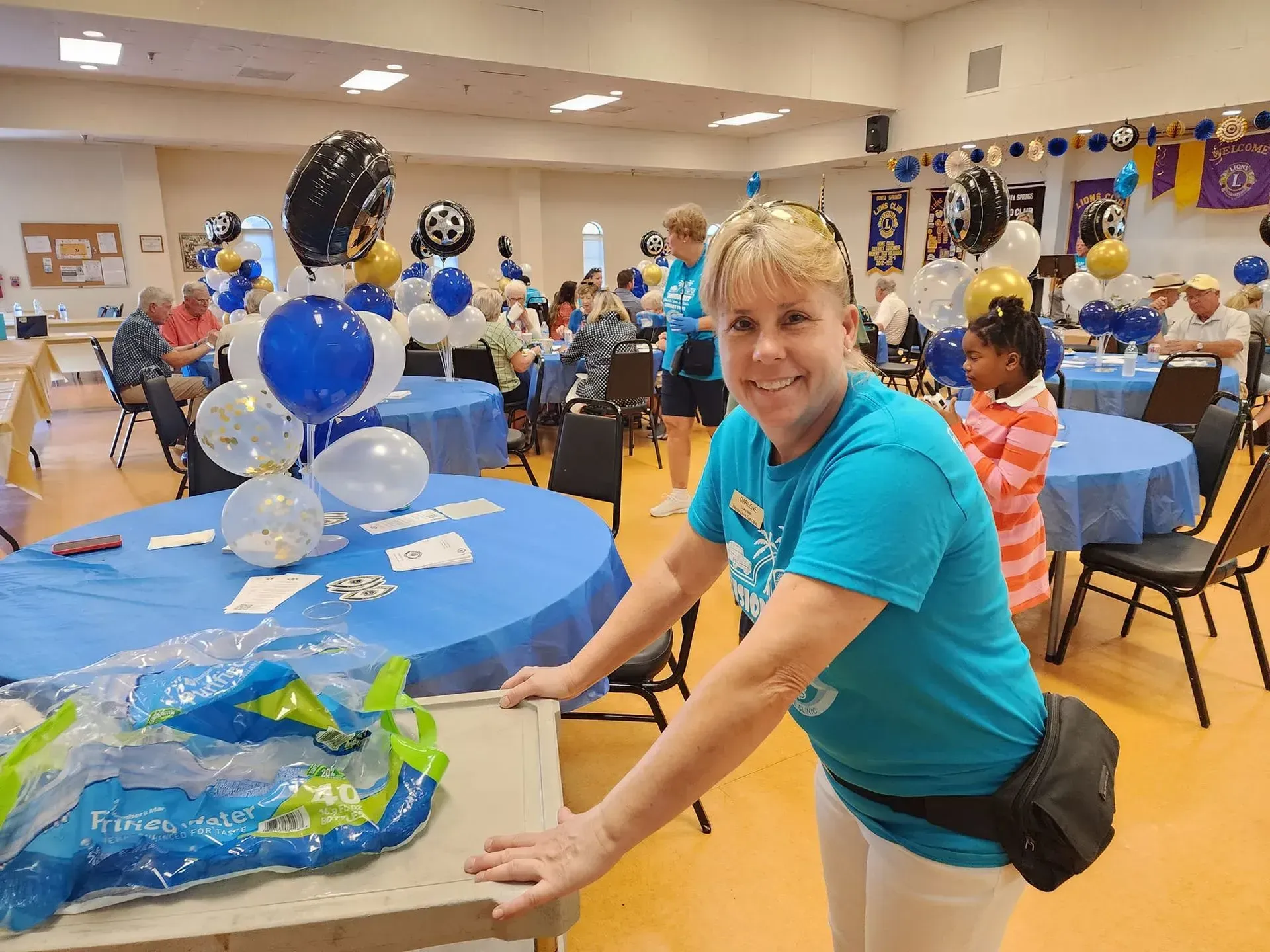 Woman in blue shirt smiles at the camera, standing by a decorated table in a community event space.