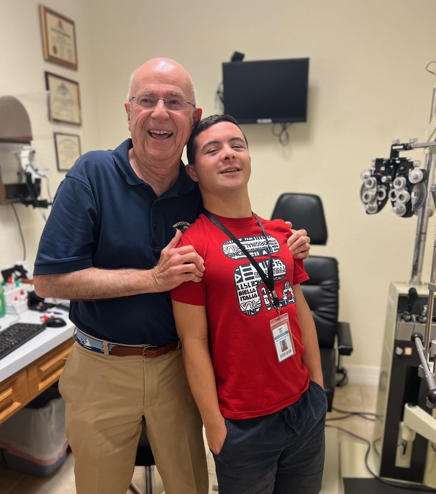 A man and a younger person pose in an eye doctor's office. Both smile, man's arms around the younger person.