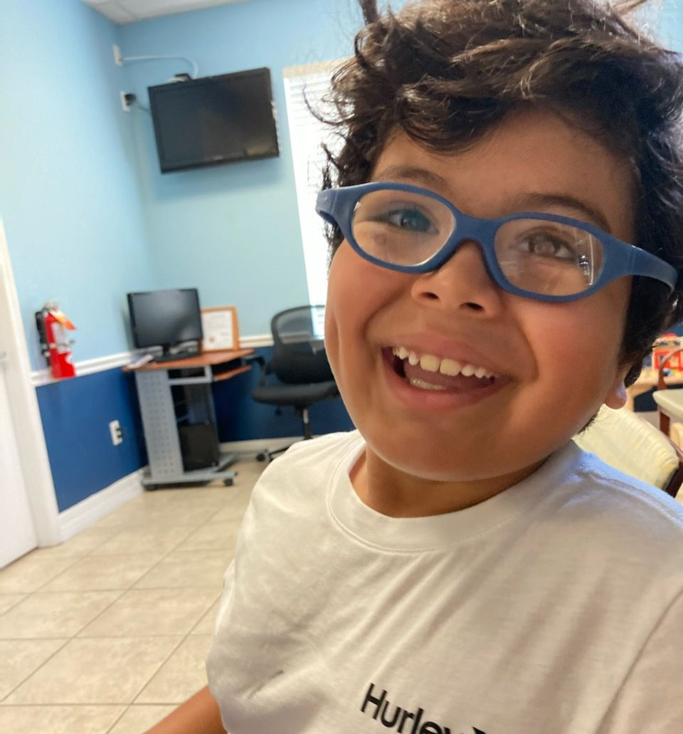 Boy with blue glasses smiles widely in a room with a computer and fire extinguisher.