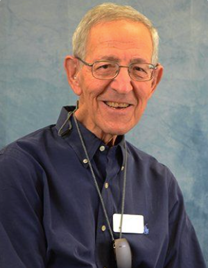Man wearing glasses and a blue shirt smiles at the camera, with a neck lanyard and a hearing aid.