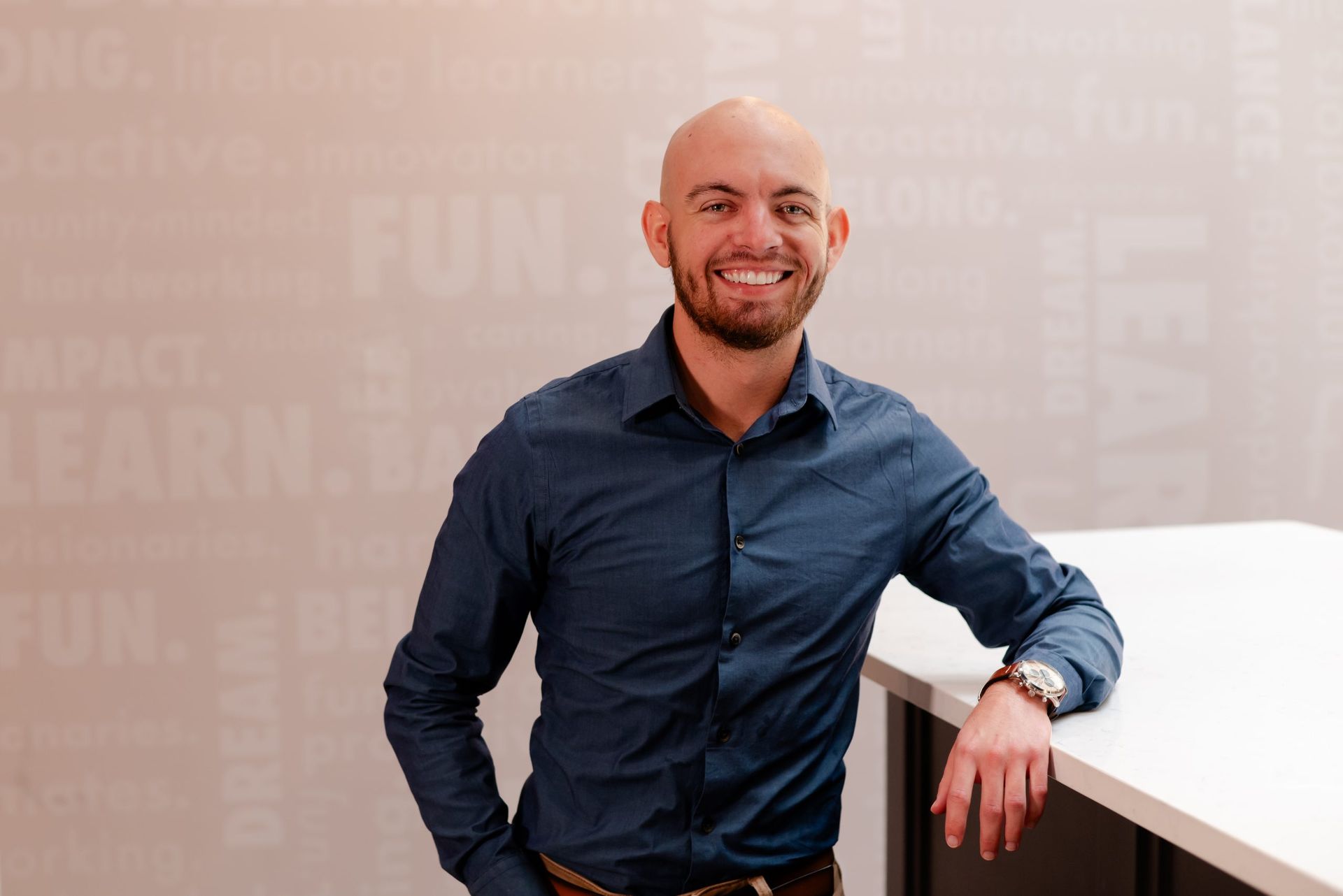 Bald man with a beard leans on a white table, smiling in front of a wall with words.