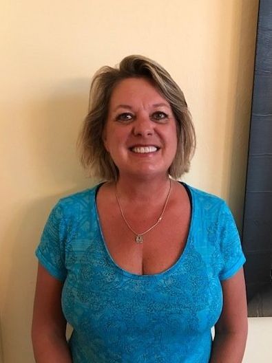 Woman with blonde hair, smiling, wearing a blue patterned top and a necklace, against a light-yellow wall.
