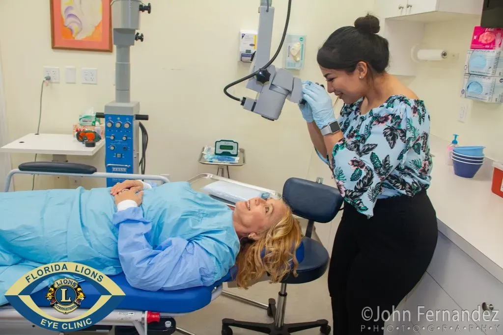 Woman in surgical gown prepares for eye exam in a clinic. Staff member adjusts equipment.
