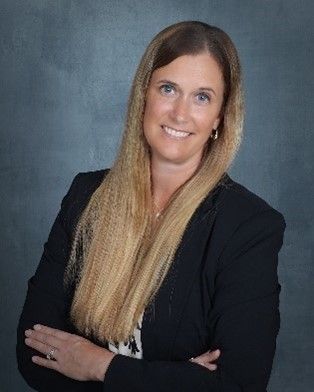 Woman in a black blazer smiles with arms crossed, against a dark blue background.