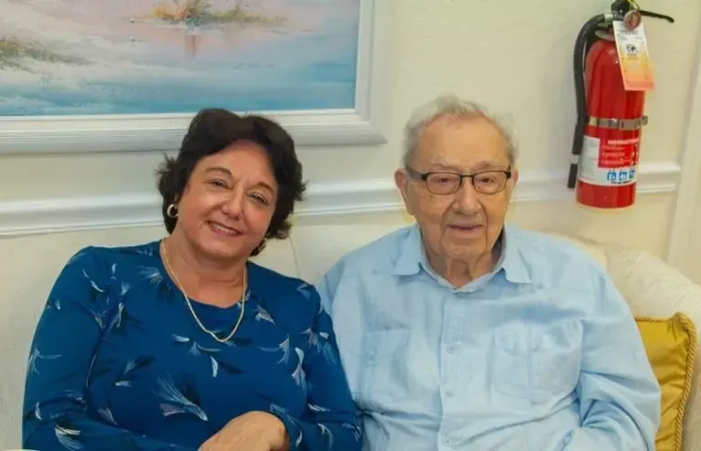 Woman and older man sit close together on a cream sofa, smiling. Blue and gold accents, fire extinguisher visible.