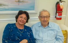 Woman and man smiling, seated on a couch in front of a painting, with a fire extinguisher visible.