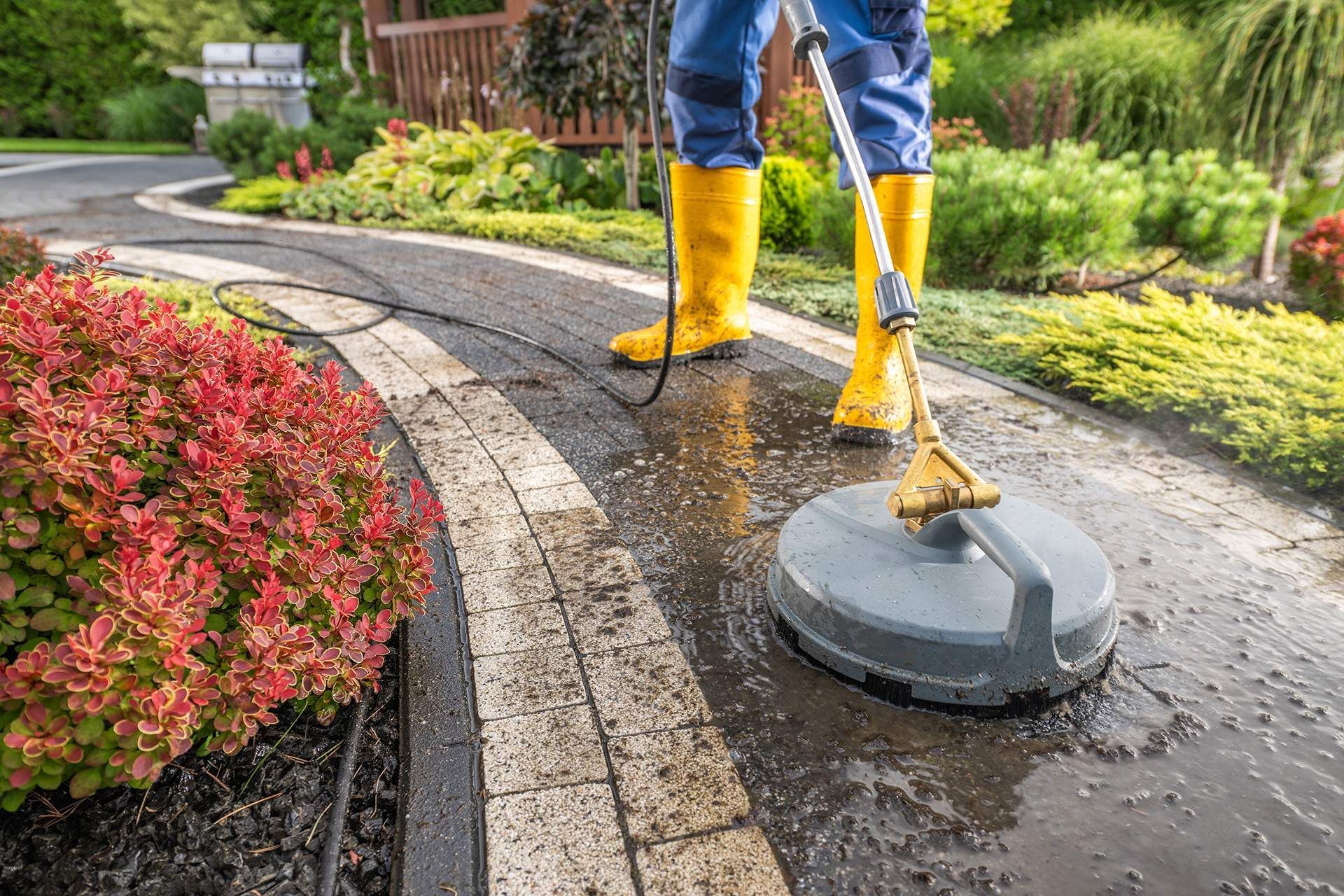 Person in yellow boots power washing a brick pathway in a garden.