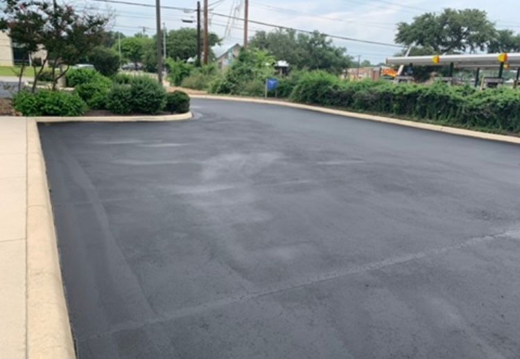 Newly paved asphalt driveway with lush green bushes and trees lining the edges.