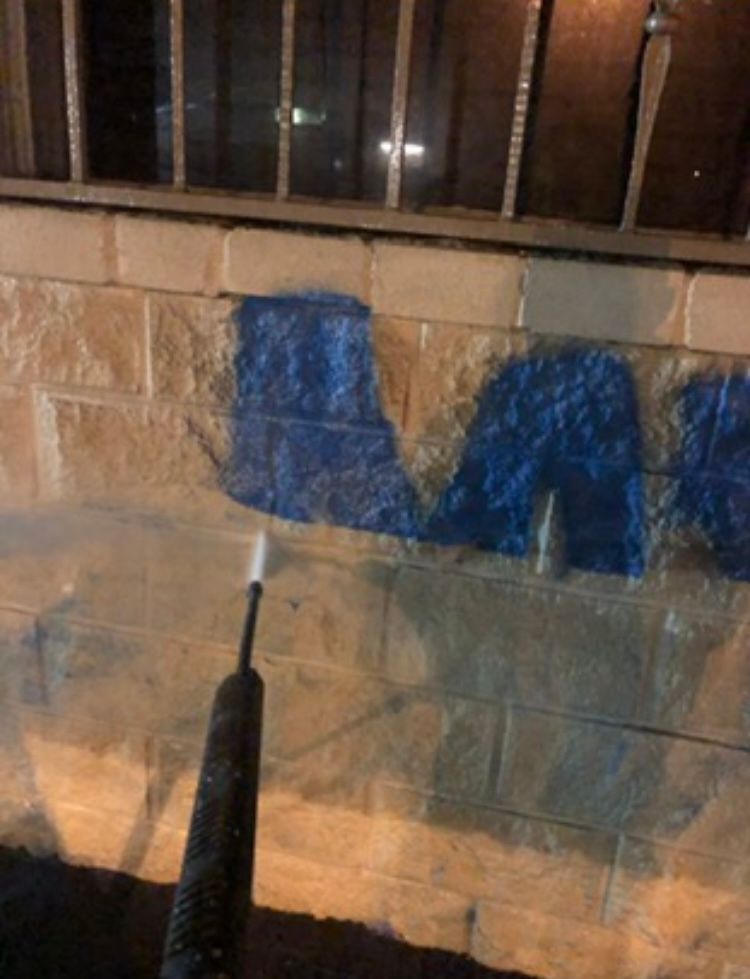 A person using a pressure washer to remove blue graffiti from a tan brick wall outside at night.