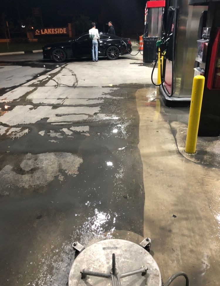 Gas station floor being cleaned with a power washer. Two black cars and people are in the background.