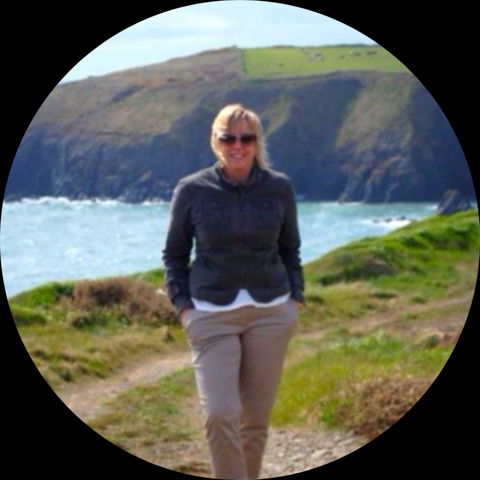 Woman in sunglasses standing on a path near a coastline. She wears a gray jacket and tan pants. Blue water and green hills are in the background.