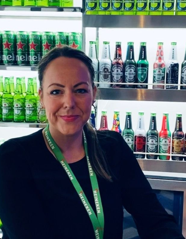 Woman with ponytail wearing a Heineken lanyard smiles in front of a Heineken display of cans and bottles.