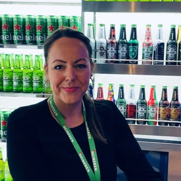 Woman wearing a Heineken lanyard stands in front of a refrigerator filled with beer bottles and cans.