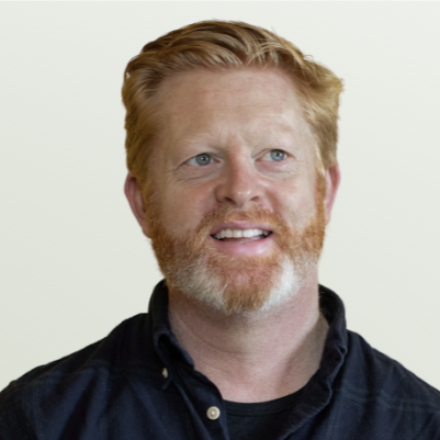 Man with red hair and beard, wearing a dark shirt, smiling with a neutral background.