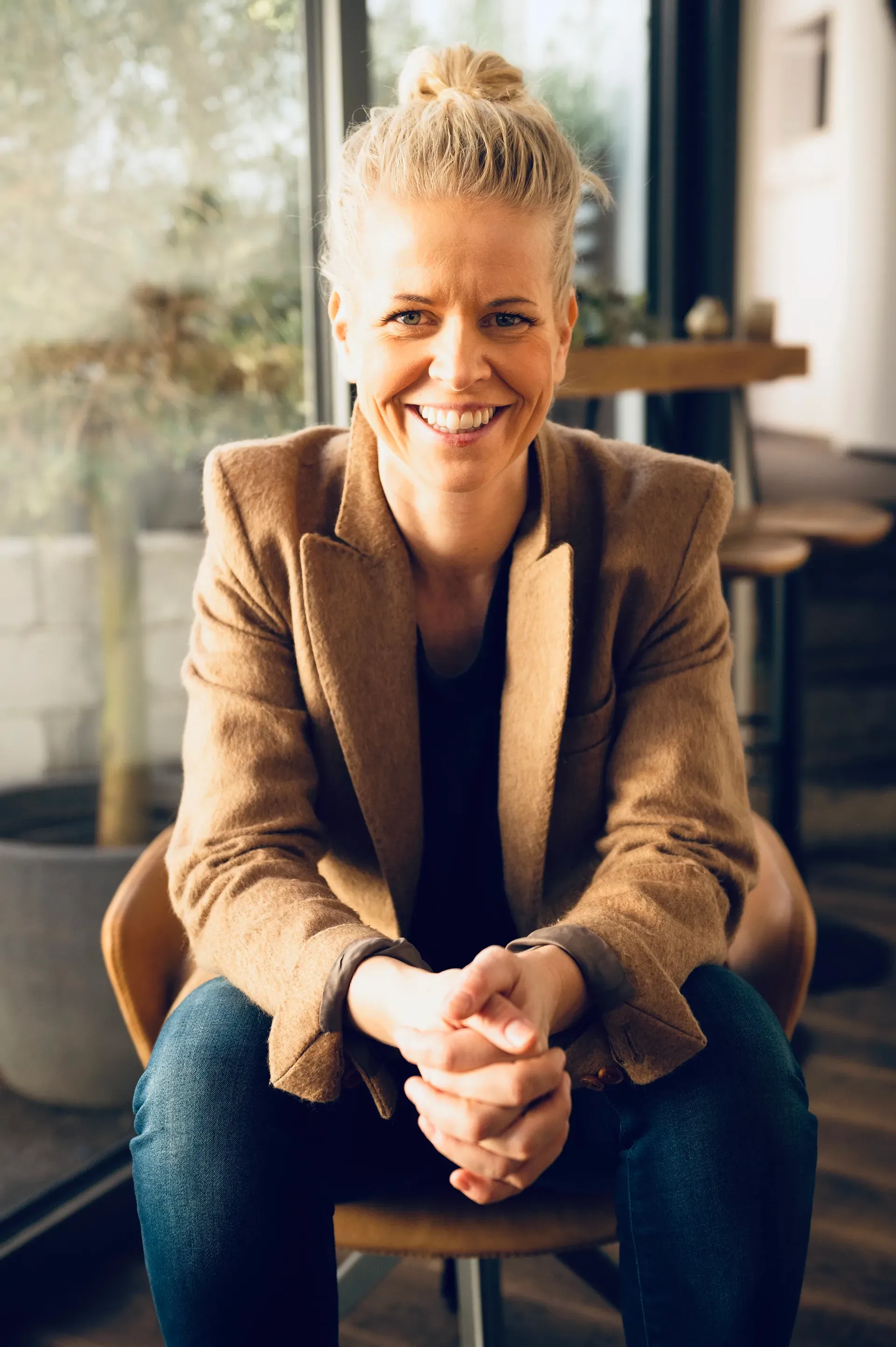 Woman with blonde hair in a bun, wearing a yellow blazer, smiling, seated at a table.