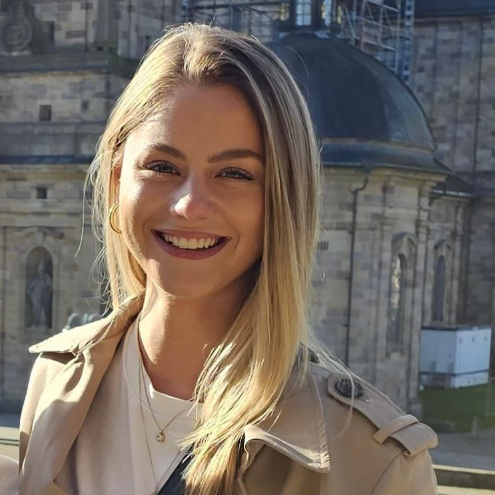 Woman with short blonde hair smiles, wearing an orange patterned shirt, in front of a Heineken sign.