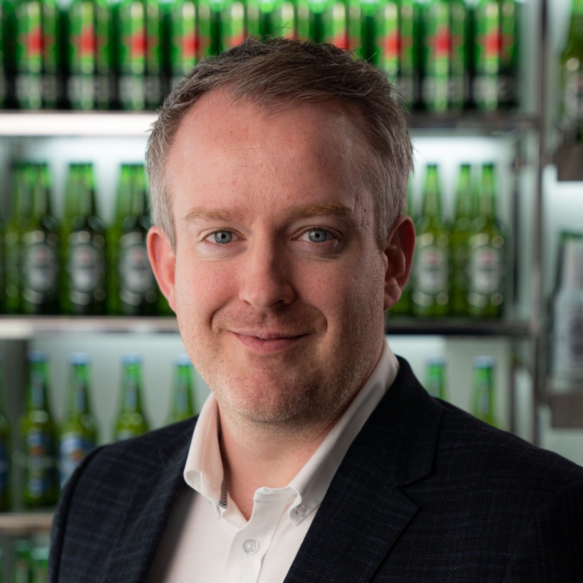 Man in a suit smiles, standing in front of shelves filled with green Heineken bottles.