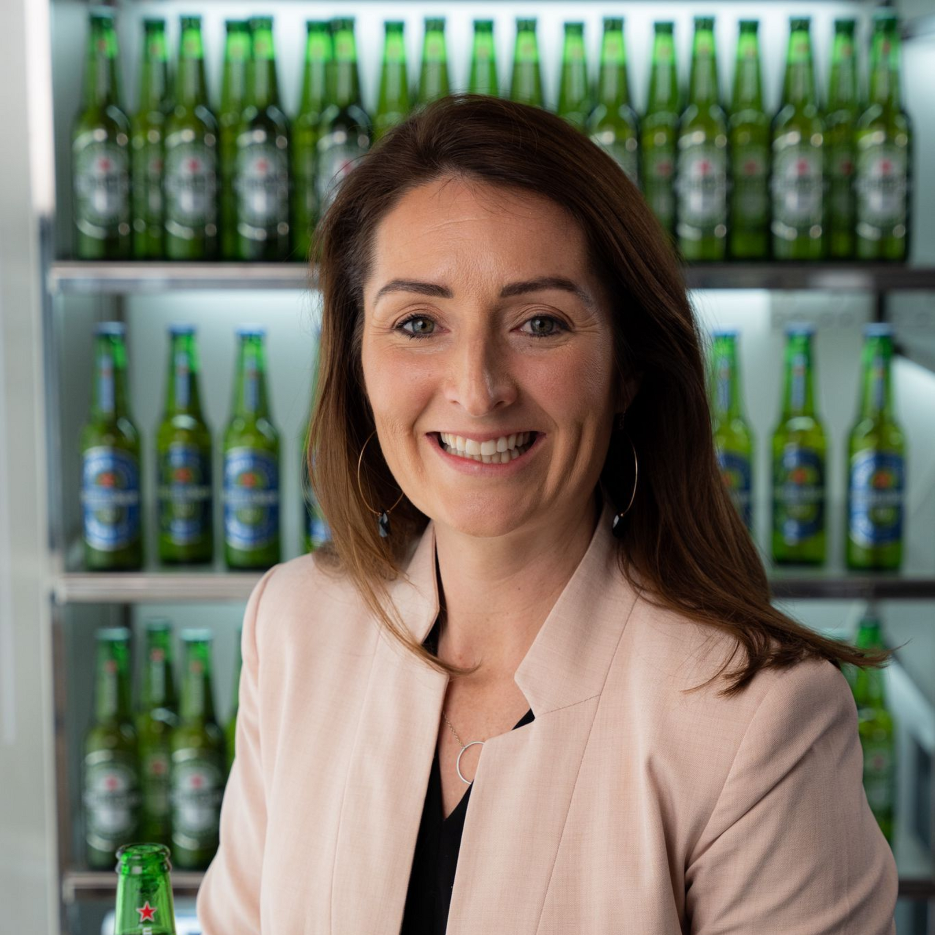 Woman in a blazer smiles in front of shelves of Heineken beer bottles.